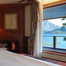 Interior view of room with interior fireplace, large window overlooking terrace, lake and mountains.