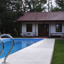 Swimming pool with small grass garden. In the background there is a colonial style construction. With wooden gates and windows. White walls and burgundy metal roof surrounded by trees.