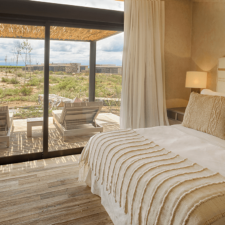 Double bed room in cream colors and adorned with fabrics and textures woven with natural wool. Overhead table and TV built into the wall. Through the window you can see a terrace and the rugged landscape.