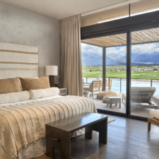 Room with a large bed with rustic details in its woven fabrics in earth colors. Warm floors and walls rich in textures. large window overlooking the terrace and in the background a beautiful landscape with river, grassland and mountains.